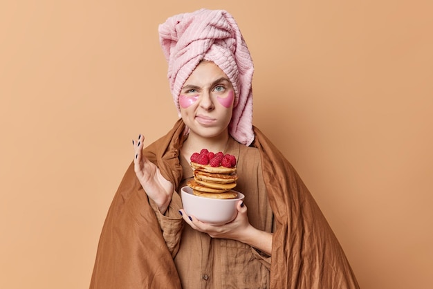 Displeased woman going to break diet and eat delicious pancakes with syrup for breakfast frowns face poses under blanket dressed in nightwear wears bath towel on head isolated over beige wall