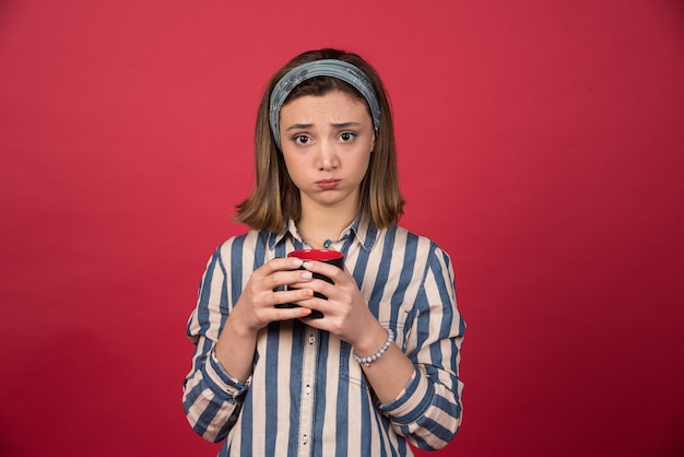 Displeased woman holding cup of coffee on red wall