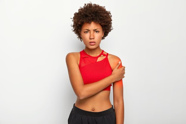 Displeased woman with slim figure, dressed in red top and trousers, touches shoulder, suffers from pain, being injured