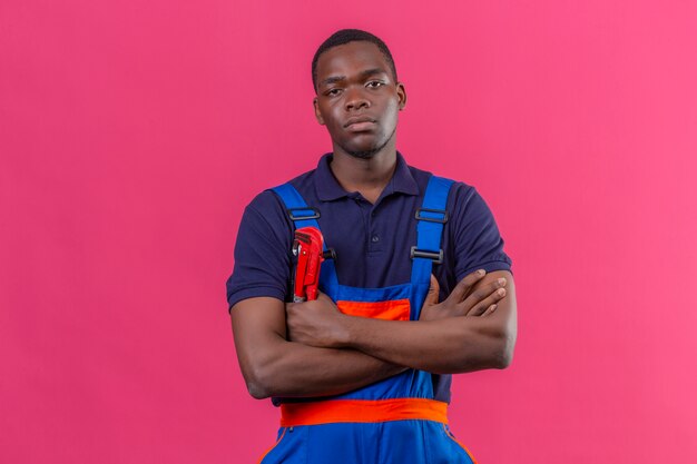 Недовольный молодой афро-американский строитель в строительной форме и кепке, стоящий со скрещенными на груди руками с несчастным лицом на розовом