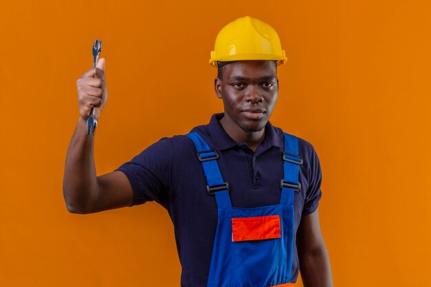 Displeased young african american builder man wearing construction uniform and safety helmet holding wrench in raised hand with angry expression standing on orange