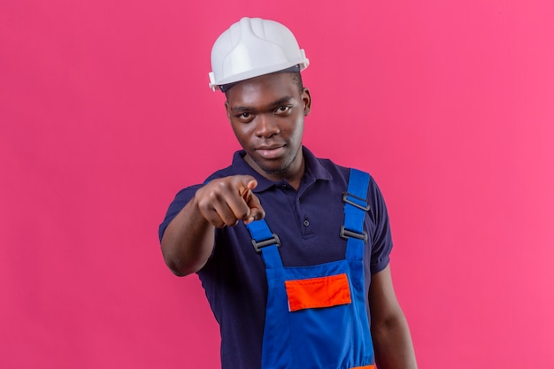 Displeased young african american builder man wearing construction uniform and safety helmet pointing finger with serious expression on face standing on pink