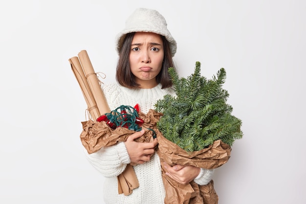 不機嫌な若いアジア人女性は顔を眉をひそめ、唇は不満を感じますトウヒの枝の花束を持っていますロール紙と装飾用のレトロな花輪はパナマと白いニットのセーターを着ています