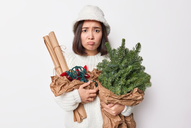 Displeased young Asian woman frowns face and purses lips feels discontent holds bouquet of spruce branches rolled paper and retro garland for decoration wears panama and white knitted sweater