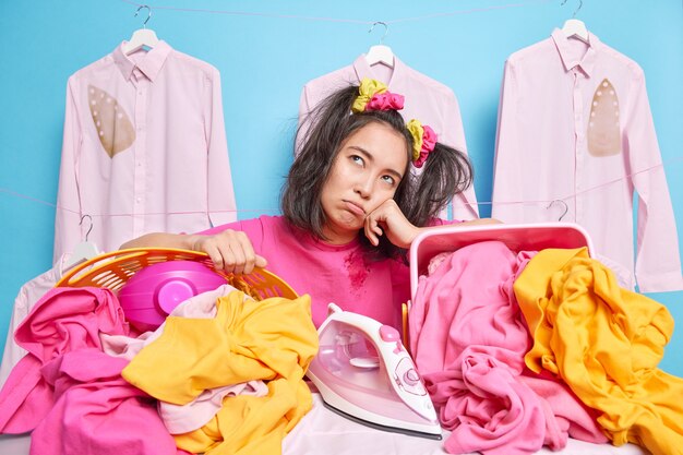 Displeased young Asian woman leans at laundry baskets feels fatigue after doing housework has tired expression
