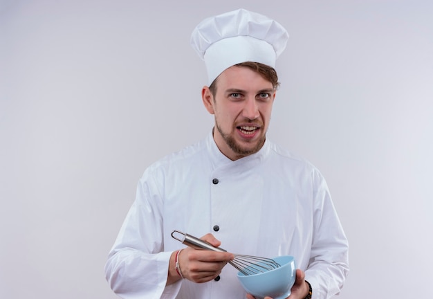 A displeased young bearded chef man wearing white cooker uniform and hat holding blue bowl with mixer spoon while looking on a white wall