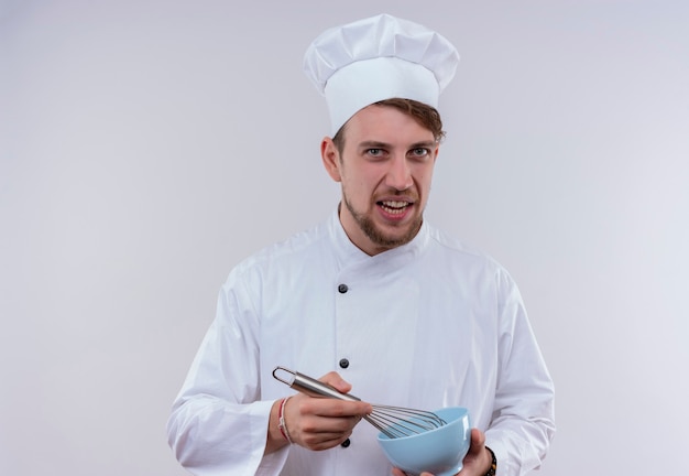 흰 벽에보고있는 동안 믹서 스푼으로 파란색 그릇을 들고 흰색 밥솥 유니폼과 모자를 쓰고 불쾌한 젊은 수염 난 요리사 남자