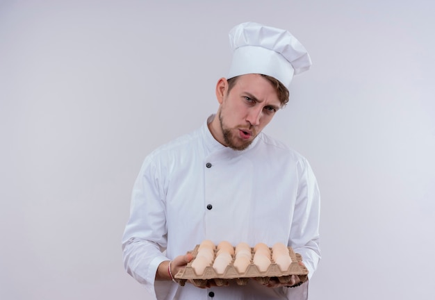 A displeased young bearded chef man wearing white cooker uniform and hat holding a carton of eggs while looking on a white wall