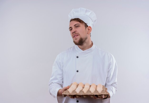 A displeased young bearded male cooker wearing white cooker uniform and hat holding a carton of eggs while looking on a white wall