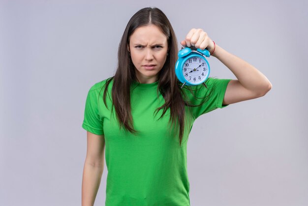 Displeased young beautiful girl wearing green t-shirt holding alarm clock  with frowning face standing over isolated white space