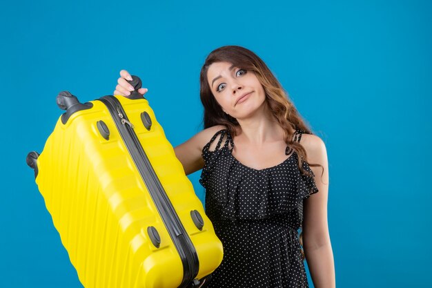 Displeased young beautiful traveler girl in dress in polka dot holding suitcase  with confuse expression on face standing over blue space