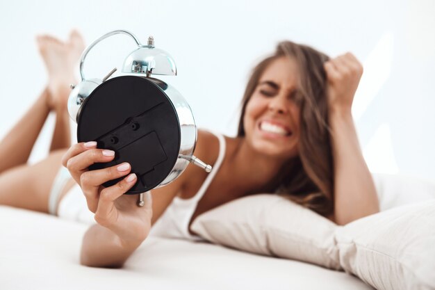 Displeased young beautiful woman lying on bed, looking at alarm-clock