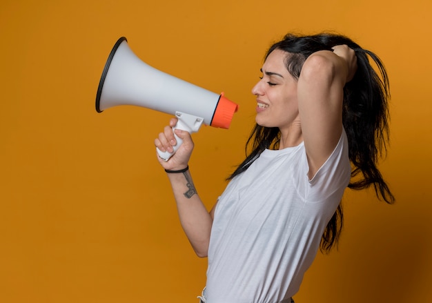 Displeased young brunette caucasian girl stands sideways and lifts hair holding loud speaker isolated on orange wall