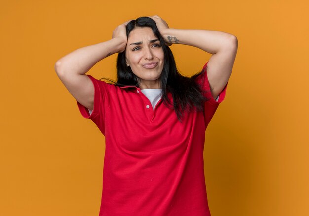 Displeased young brunette caucasian girl wearing red shirt holds head isolated on orange wall