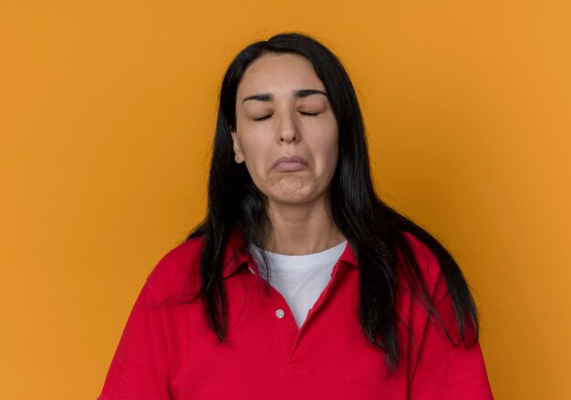 Displeased young brunette caucasian girl wearing red shirt stands with closed eyes isolated on orange wall