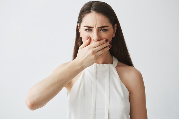 Displeased young brunette girl covering mouth with hand over white backround.