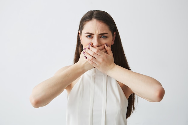 Displeased young brunette girl covering mouth with hand over white backround.