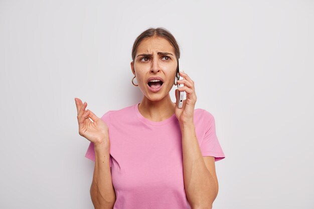 不機嫌な若いブルネットの女性は、不快な顔からの怒りの表情を動揺させています退屈な電話の会話困惑した表情は白い背景の上に分離されたカジュアルなピンクのTシャツを着ています