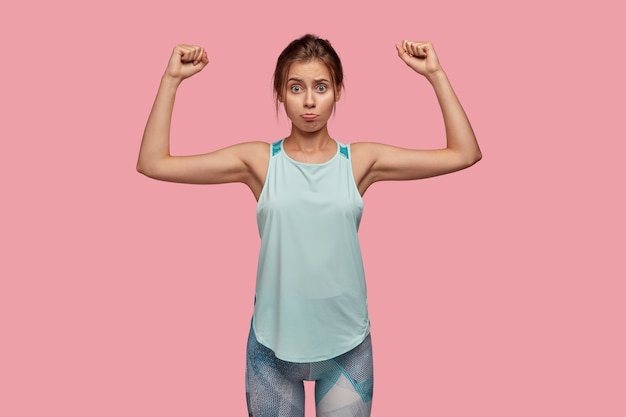 Displeased young Caucasian blue eyed woman with unhappy sullen expression, has nice perfect figure raises hands and shows muscles, wears activewear, isolated over pink wall, feels weak