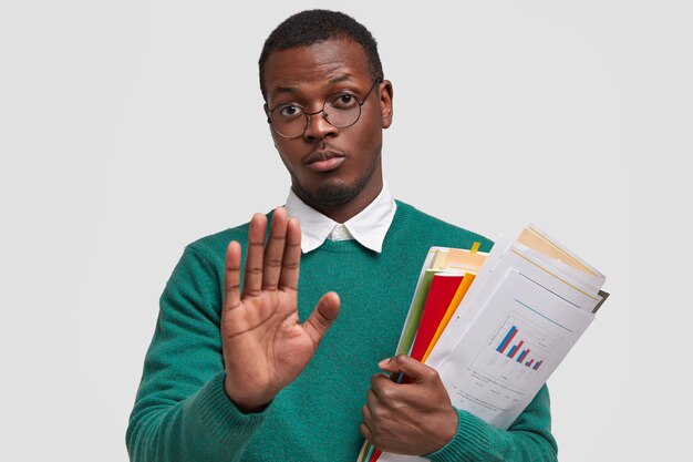 Displeased young entrepreneur with dark skin, holds documentation and notepad, demonstrates stop gesture, refuses to develop business ideas alone