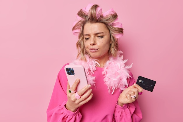 Displeased young European woman applies hair rollers checks banking account via smartphone holds credit card wears dress isolated over pink background. Online payment and purchasing concept.