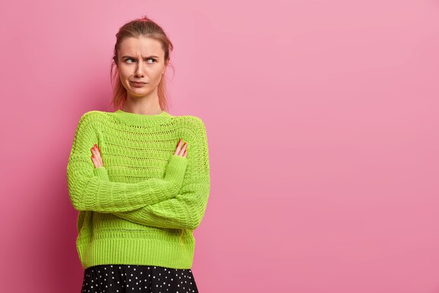 Displeased young European woman being angry, keeps arms folded, smirks face, stands in offended pose, wears green knitted sweater