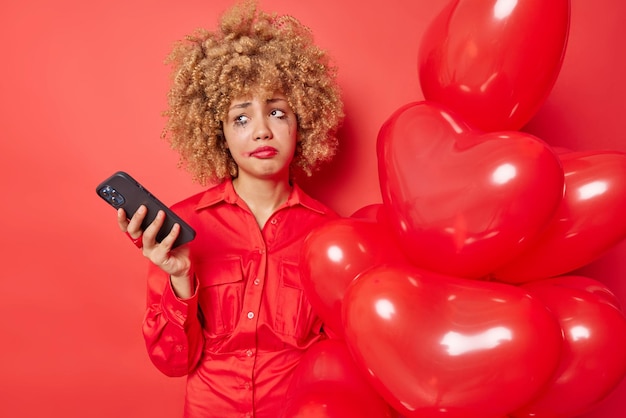 Displeased young european woman has spoiled mood waits forlover on valentines day uses mobile phone feels lonely holds bunch of heart balloons has leaked makeup isolated over red background