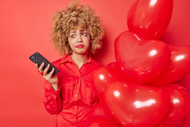 不機嫌な若いヨーロッパの女性は気分を台無しにしましたバレンタインデーに恋人を待っています携帯電話を使用して孤独を感じますハートの風船の束が赤い背景の上に分離された化粧を漏らしました
