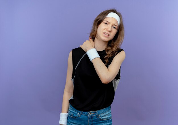 Displeased young fitness girl in black sportswear with headband touching her shoulder having pain standing over blue wall