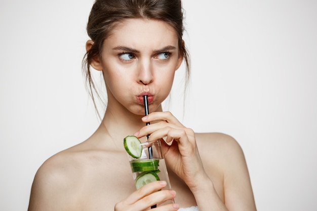 Displeased young girl drinking water with cucumber slices over white background. Beauty cosmetology and spa. Facial treatment.