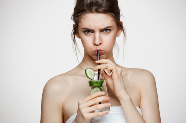 Displeased young girl looking at camera drinking water with cucumber slices over white background. Beauty cosmetology and spa. Facial treatment.