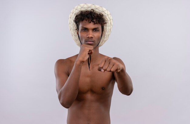 A displeased young handsome dark-skinned man with curly hair wearing sun hat showing boxing moves while  