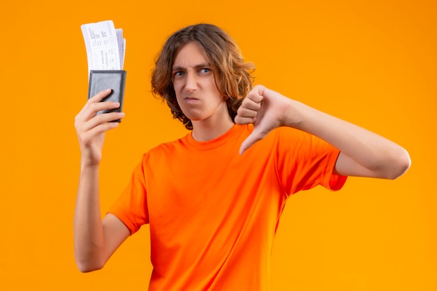 Displeased young handsome guy in orange t-shirt holding air tickets showing thumbs down standing over orange background