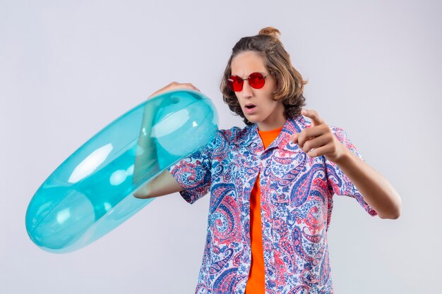 Displeased young handsome guy wearing red sunglasses holding inflatable ring pointing to camera with angry expression standing 