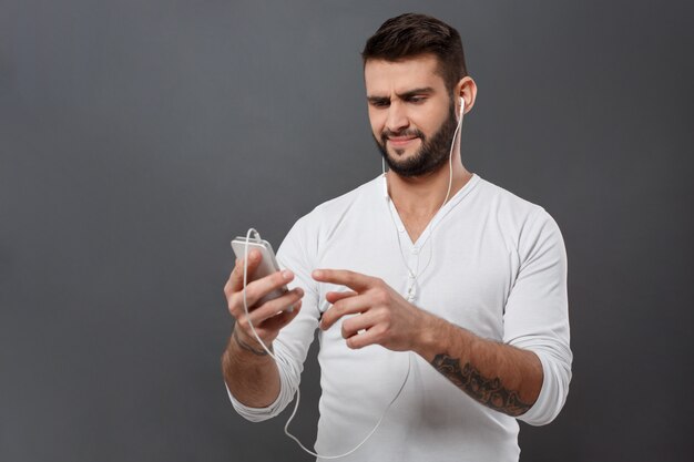 Displeased young handsome man looking at phone over grey wall