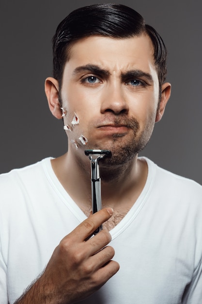 Displeased young handsome man shaving on grey