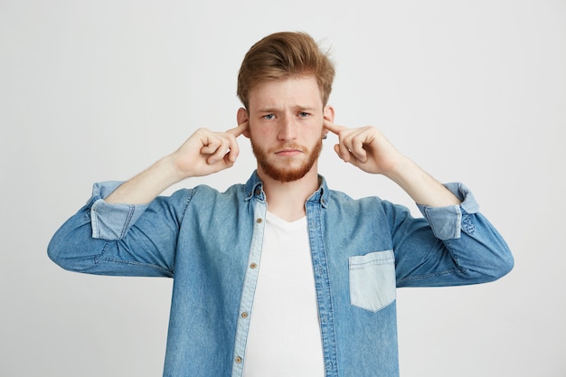 Displeased young handsome man with beard frowning closing ears.