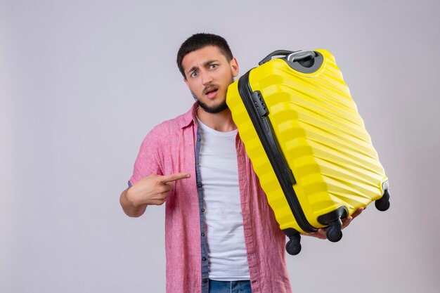 Displeased young handsome traveler guy holding suitcase pointing with finger to it  with confuse expression on face standing 