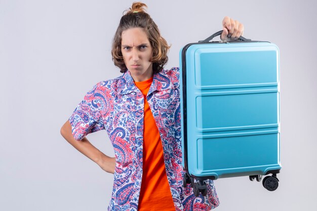 Displeased young handsome traveler guy holding suitcase on shoulder  frowning with anger standing 