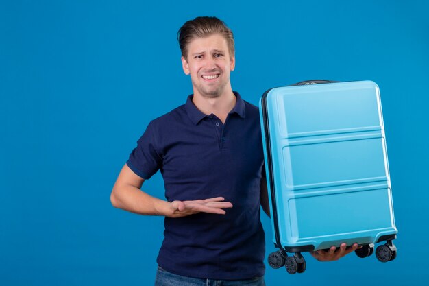 Displeased young handsome traveler man holding suitcase presenting with arm of his hand with confuse expression on face standing over blue space