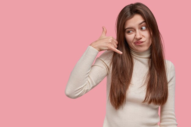 Displeased young lovely lady gestures indoor, makes call sign with hand