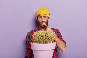 Бесплатное фото Недовольный молодой человек уколол палец от шипа кактуса, стоит возле растения в горшке, носит желтую шляпу
