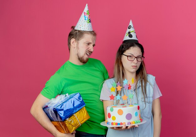 파티 모자를 쓰고 불쾌한 젊은 남자가 파티 모자를 쓰고 분홍색 벽에 고립 된 생일 케이크를 들고 짜증이 어린 소녀를보고 선물 상자를 보유하고 있습니다.