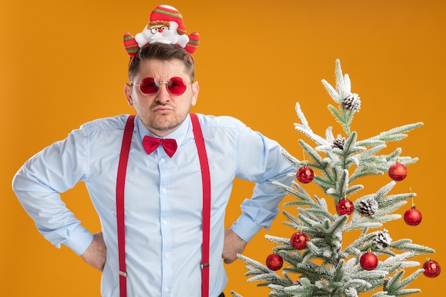오렌지 배경 위에 성난 얼굴로 카메라를보고 크리스마스 트리 옆에 서있는 산타와 빨간 안경 테두리에 멜빵 나비 넥타이를 착용하는 불쾌한 젊은 남자