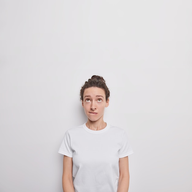 Displeased young millennial woman bites lips focused above listens something with attentive gaze upwards poses against white wall