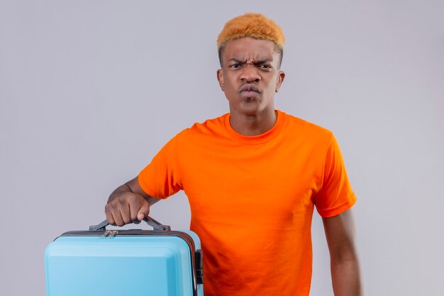 Displeased young traveler boy wearing orange t-shirt holding suitcase with frowning face standing over white wall