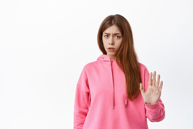 Displeased young woman blocking offer, refusing something bad, showing stop block gesture, rejecting, standing over white wall