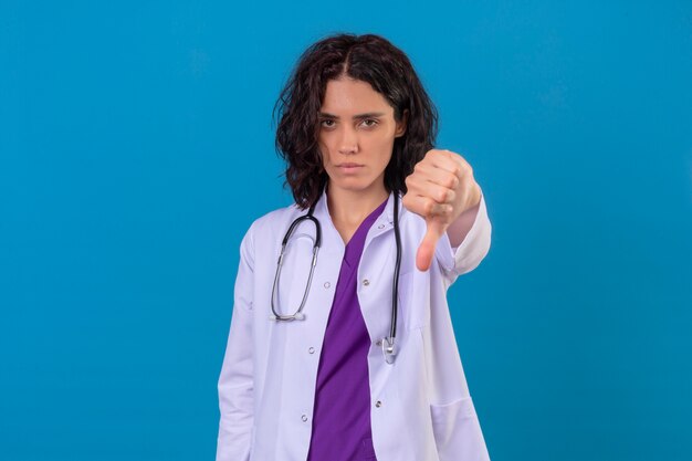 Displeased young woman doctor wearing white coat with stethoscope showing thumb down standing on isolated blue