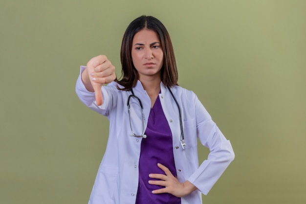 Foto gratuita medico dispiaciuto della giovane donna che indossa camice bianco e con lo stetoscopio che mostra i pollici giù in piedi sul verde isolato