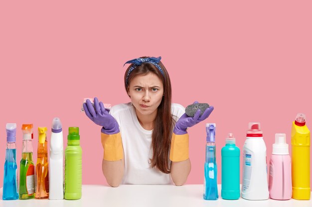 Displeased young woman frowns face in discontent, carries sponges, wears white casual t shirt, rubber gloves, looks angrily in camera